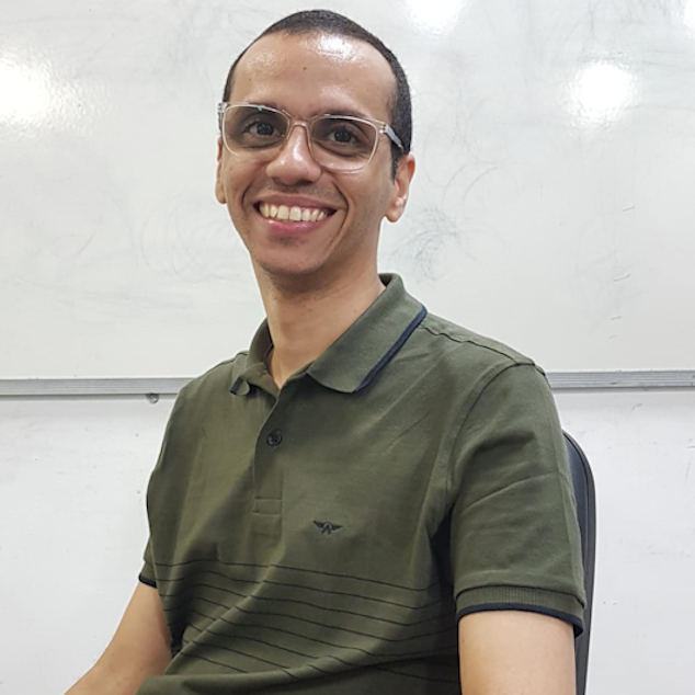 Jovem; de cor parda; cabelos pretos rente a cabeça; olhos pequenos e pretos; rosto ovalado; usa óculos de grau grande; lábios finos; nariz pequeno fino. Usa uma blusa verde musgo e está diante de uma parede branca, com enquadramento abaixo dos ombros.