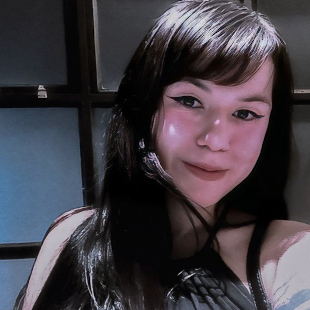 Mulher branca, de cabelo preto com franja, usando um vestido preto, sorrindo. Ao fundo uma porta de vidro com vigas de madeira.