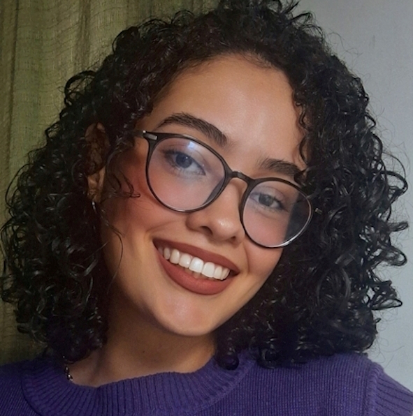 Jovem de pele parda com cabelos cacheados na cor marrom escuro e olhos pretos com óculos de armação preta. Está sorrindo diante de uma parede branca com cortinas verdes claro, veste uma blusa roxa e usa um batom marrom