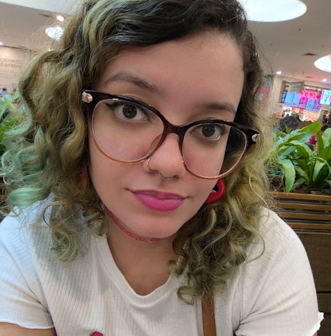 Jovem de pele branca, tem cabelos ondulados e esverdeados na altura dos ombros e olhos castanhos escuros. Usa óculos de armação marrom para preto, batom rosado e blusa branca. Está sentada diante de algumas plantas.