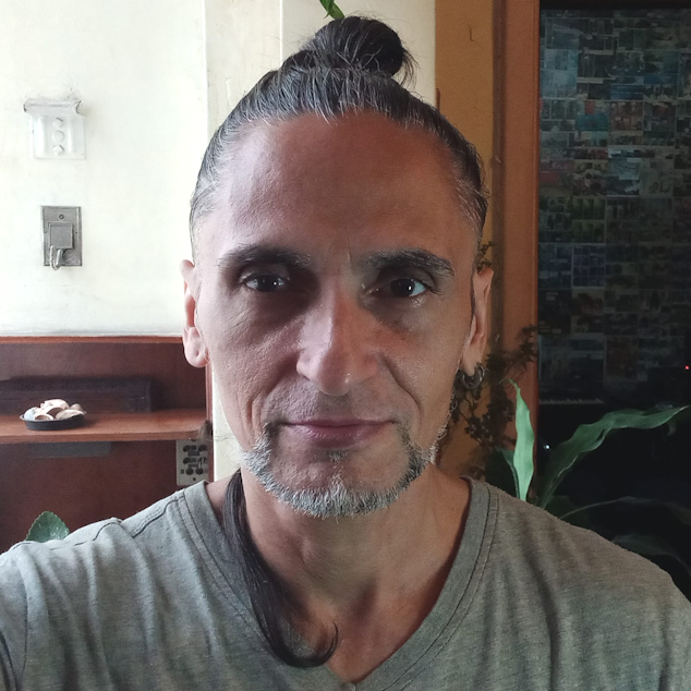 Foto de um homem adulto, de pele parda clara, cabelos lisos compridos castanhos presos em coque e olhos castanhos. Sua barba em cavanhaque é parcialmente grisalha. Ele está com um leve sorriso. Veste uma camisa cinza com gola em V. Está em pé. Ao fundo, há uma parede amarela, uma prateleira de madeira e algumas plantas. A luz do ambiente é matinal, suave e acolhedora.